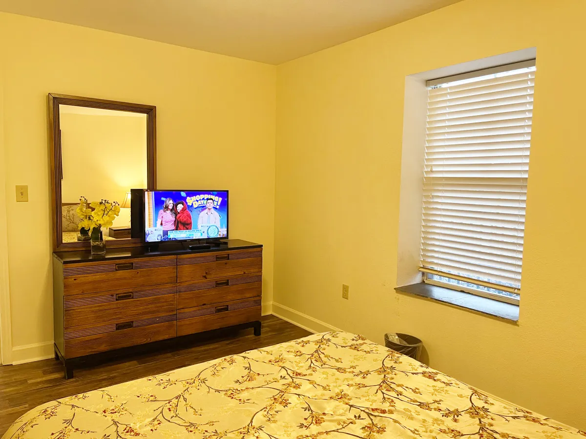 A cozy bedroom featuring a queen bed adorned with a floral comforter, a wooden dresser topped with a TV, and a window with blinds letting in gentle daylight. The space feels comfortable and inviting, ideal for restful privacy.
