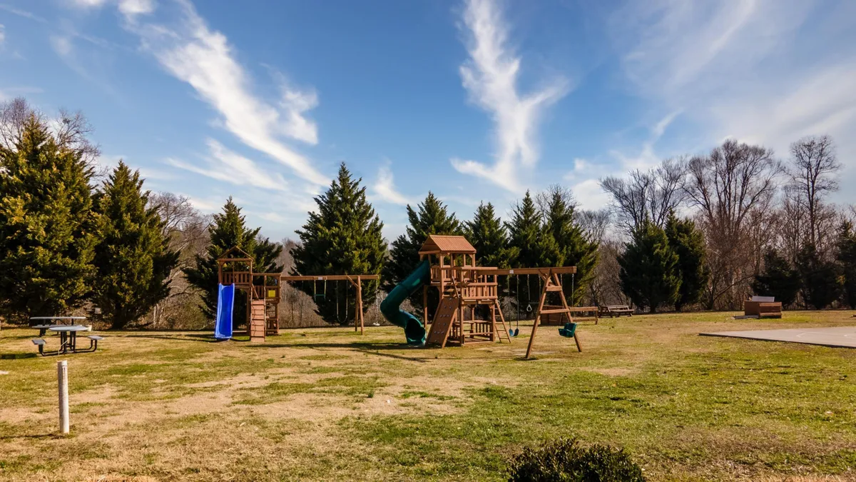 A joyful playground scene with a colorful slide, swings, and picnic tables set against green grass and trees. The area is bustling with opportunities for children’s play and family picnics.