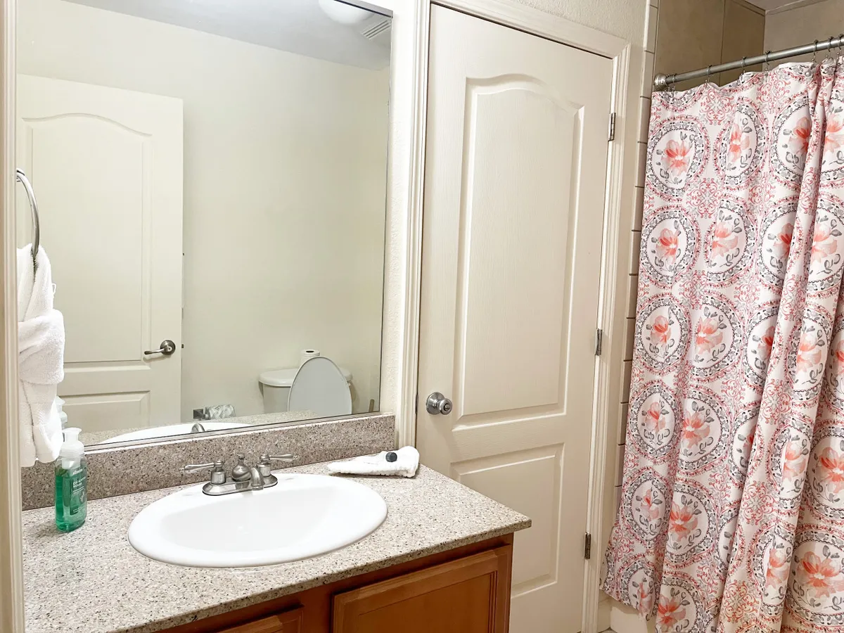 A calm bathroom scene with a vanity, sink, decorative shower curtain, and a mirror reflecting the clean, airy space. Soft lighting hints at the presence of fresh linens and towels, contributing to a sense of cleanliness and well-being.
