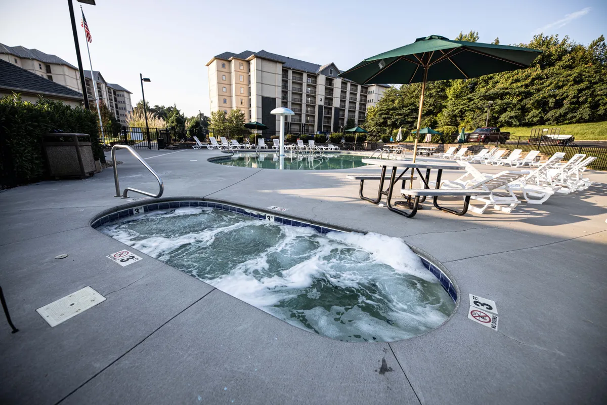 This image features a tranquil outdoor scene with a large hot tub next to a swimming pool, surrounded by lounge chairs and a shaded table. The setting suggests leisurely poolside gatherings, relaxation in the hot tub, and sociable sunbathing.