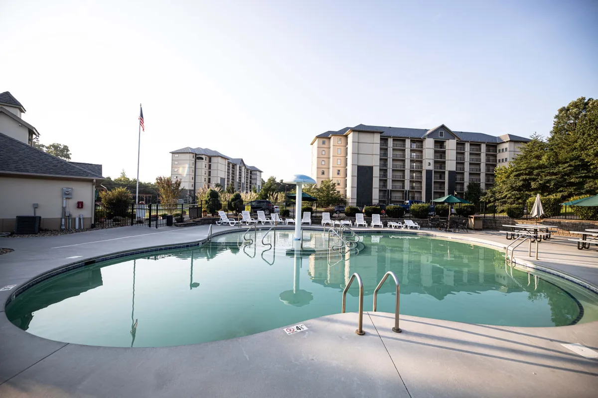 A spacious outdoor swimming pool surrounded by lounge chairs and umbrellas, with well-maintained landscaping and a flag pole visible in the background. The sunny setting invites relaxation and active play for families.