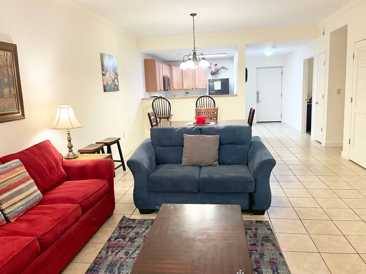 An inviting open-plan living and dining area featuring a red sofa, a blue sofa, a dining table set for six, and colorful artwork on the wall. The setup encourages communal activities, with ample seating for relaxation or conversation, and a dining space seamlessly connected to the living area.
