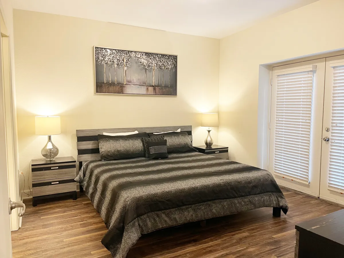 A serene master bedroom with dark bedding, decorative artwork above the bed, matching nightstands with lamps, and sliding doors leading to the outside. The space is bathed in soft natural light, enhancing its tranquil atmosphere.