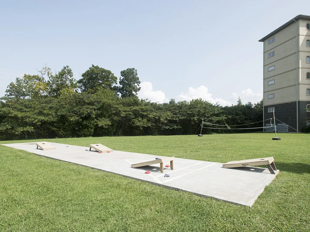 A grassy outdoor recreation area featuring cornhole boards, a volleyball net, and the building façade in the background. The scene is lively and playful, designed for group activities.