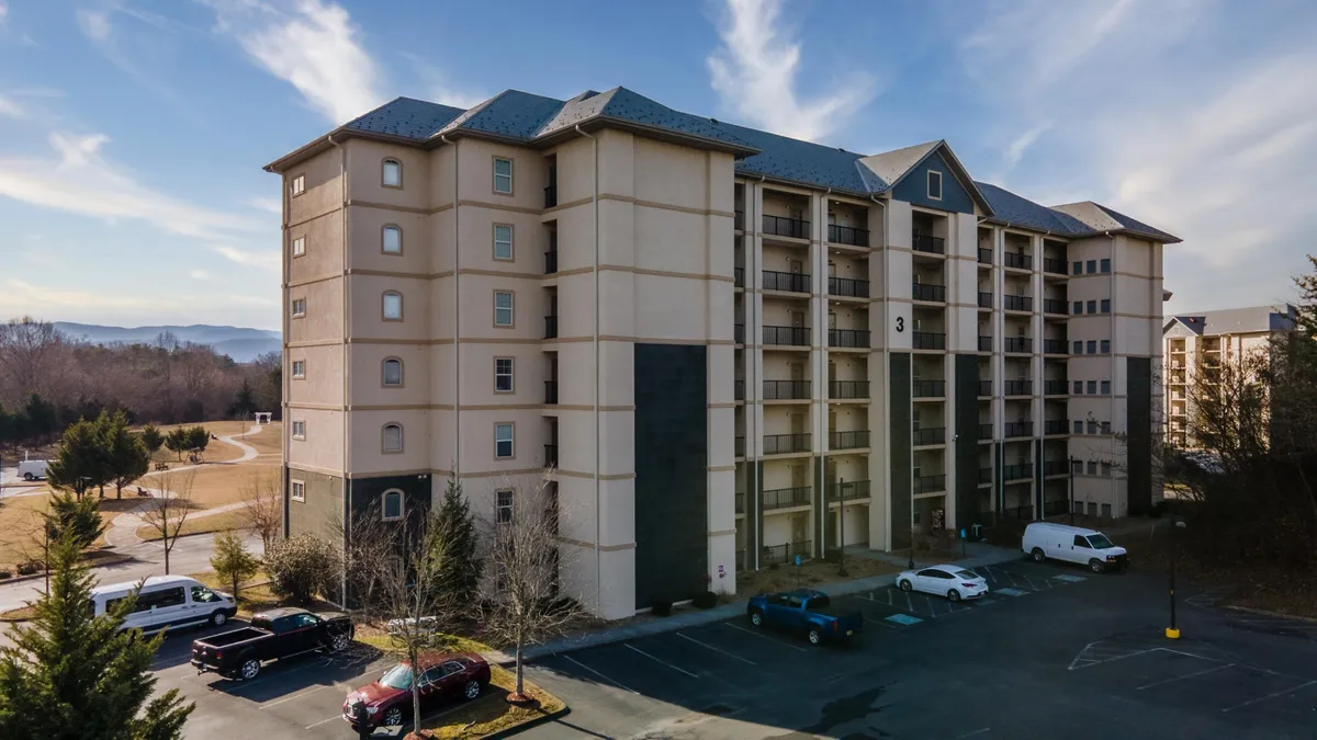 A four-story condominium building with ample parking space in front, surrounded by lush trees and a majestic mountain backdrop. The building’s ground-level entry is visibly accessible, and there are balconies for each unit, suggesting spaces for group gatherings and relaxation. The welcoming environment hints at on-site amenities and ease of access for all guests.