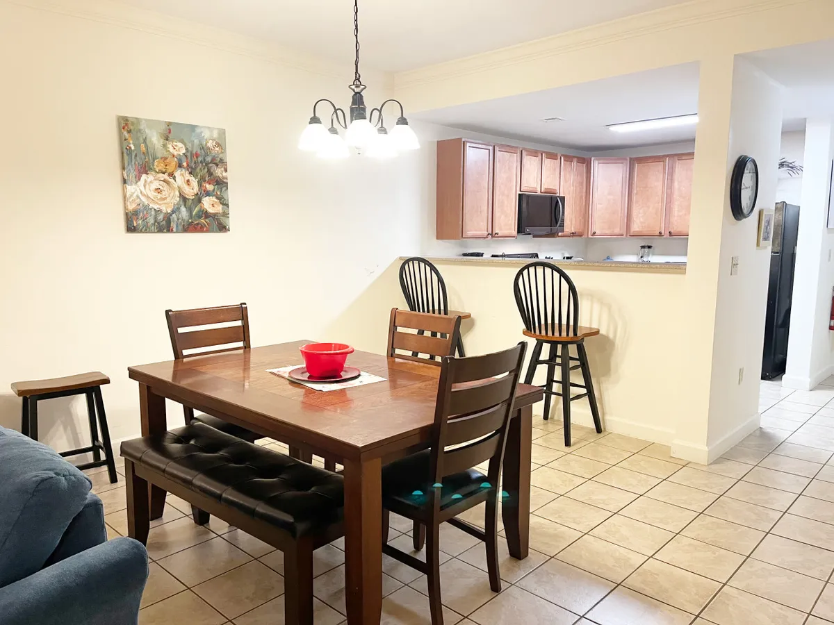 A warm, cheerful dining space with a dining table for six, a kitchen bar, floral artwork, and an elegant light fixture. The room is styled for both casual meals and festive family gatherings, reinforcing the welcoming, home-like atmosphere.