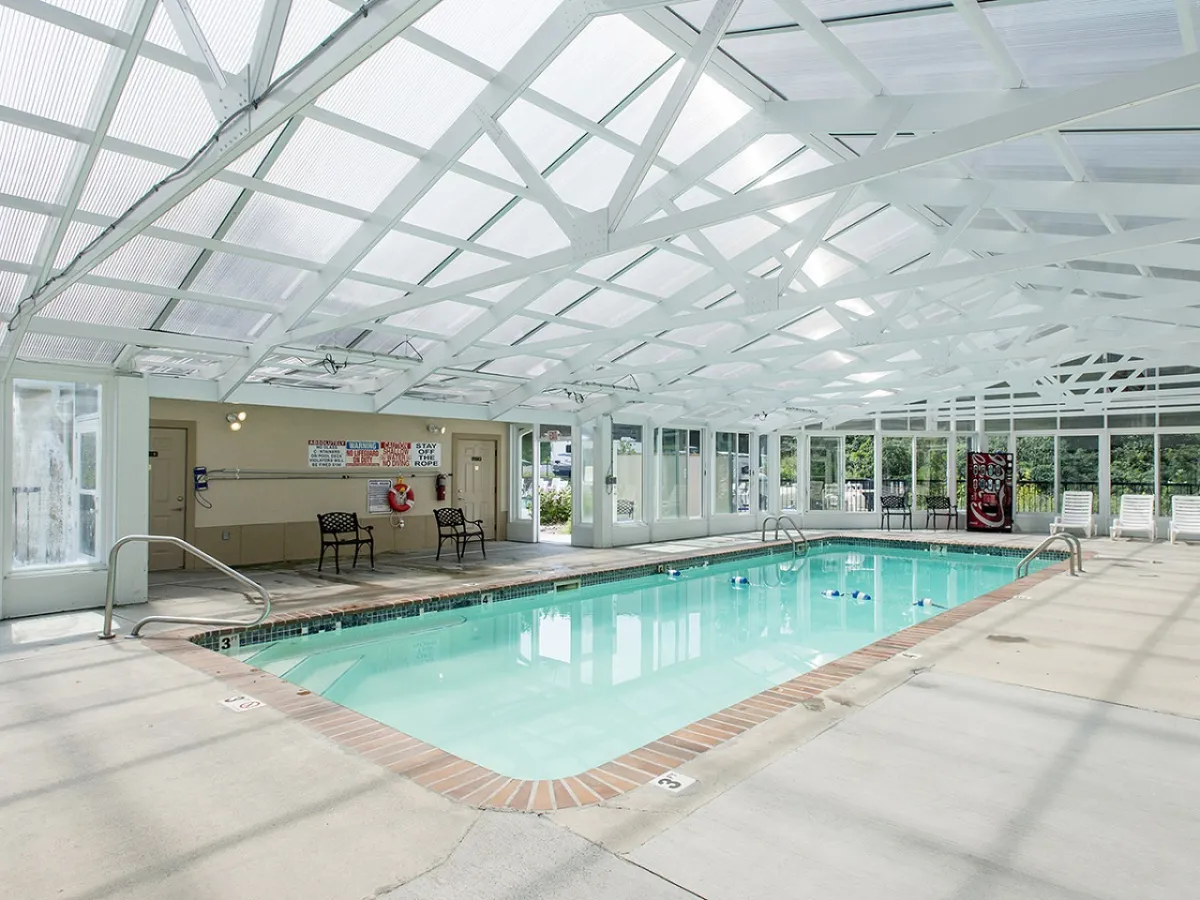 A bright, airy indoor swimming pool is shown with large glass windows letting in abundant natural light. White ceiling beams and poolside chairs add to the relaxing vibe, making it inviting for both swimming and relaxation year-round.